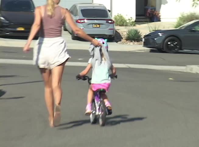 A mother runs alongside her young daughter, helping her learn to ride her bicycle on a suburban street