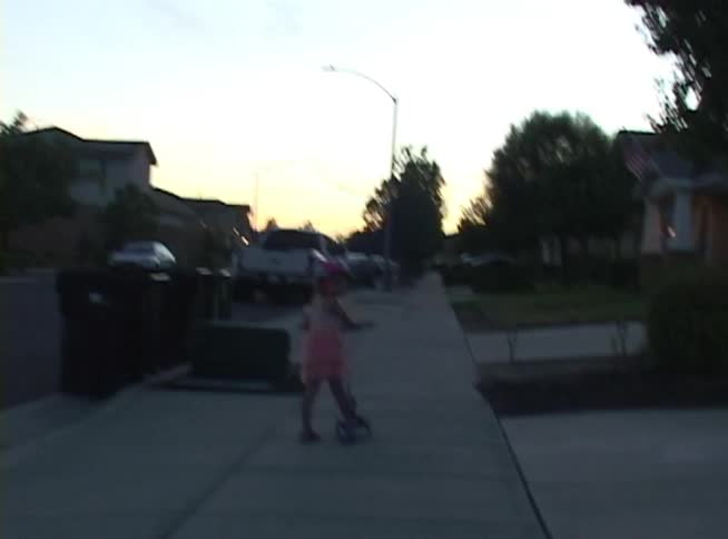 An airplane flies across the blue sky, followed by a shot of a little girl in a pink dress riding her scooter down a suburban sidewalk at dusk