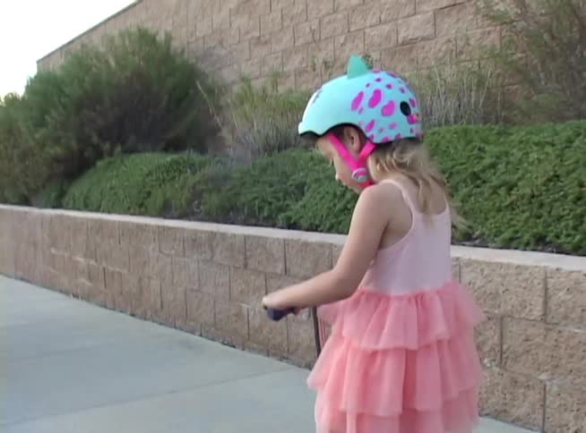 A young girl in a pink frilly dress and a novelty helmet rides her scooter slowly down a neighborhood sidewalk
