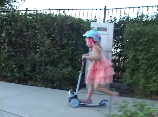 A little girl in a pink dress and helmet joyfully rides her scooter down a neighborhood sidewalk