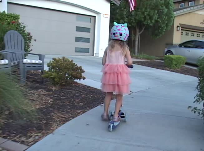 Following a young girl in a pink dress and animal helmet as she rides her scooter home down the sidewalk