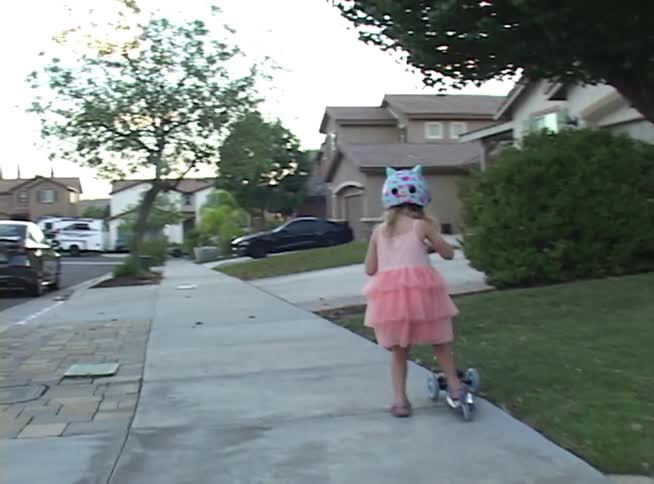 A young girl in a pink dress and helmet rides her three-wheeled scooter in slow motion down a suburban sidewalk