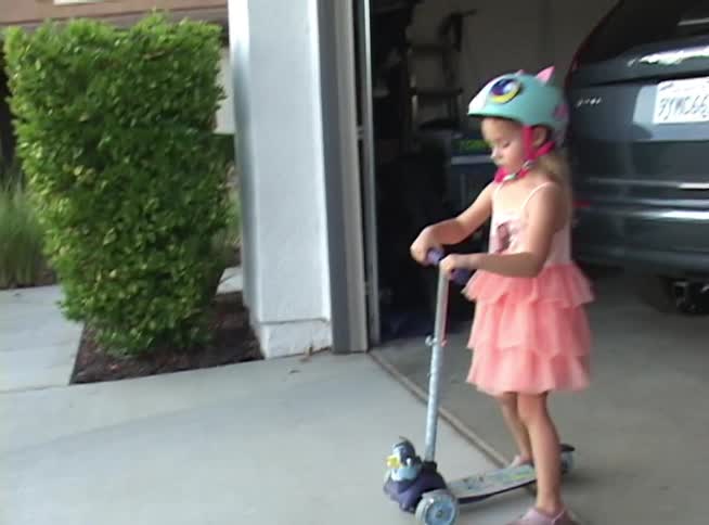 A young girl in a pink dress and colorful cat helmet gets ready to ride her scooter in the driveway