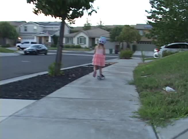 A young girl in a pink dress and helmet rides her scooter down the sidewalk in a quiet suburban neighborhood