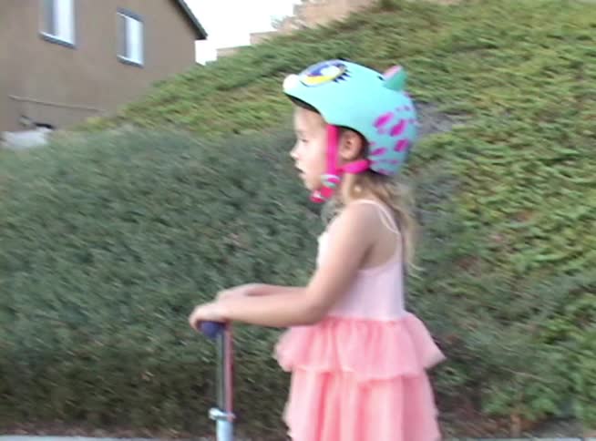 A young girl in a pink dress and a colorful helmet rides her scooter down a suburban sidewalk before stopping to think