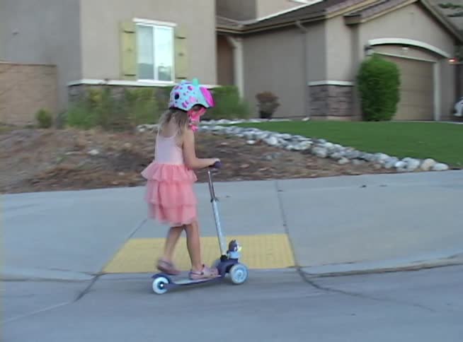 A young girl in a pink dress and helmet rides her scooter down a suburban street and onto the sidewalk