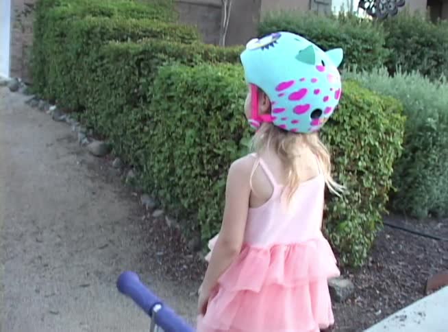 A little girl in a pink dress and helmet points at a 'No Trespassing' sign before walking down a path with her scooter