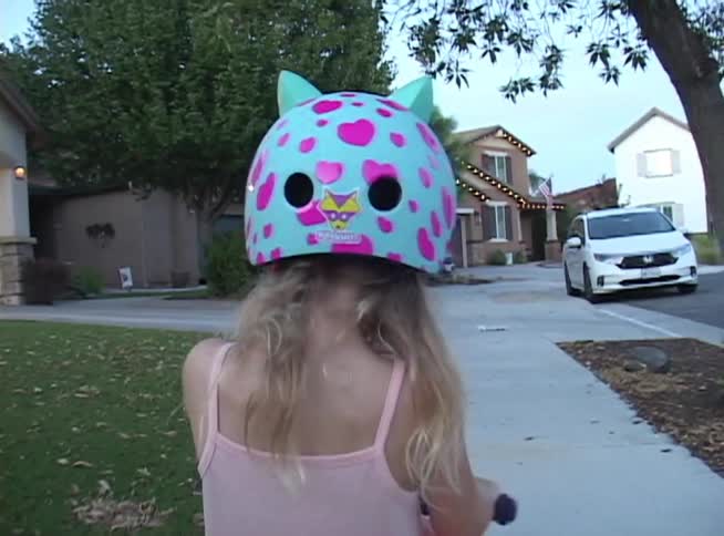 A little girl in a pink dress and a cute cat-eared helmet rides her scooter down a suburban sidewalk in the evening