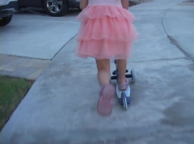 Following a young girl in a pink ruffled dress and a cute helmet as she rides her three-wheeled scooter down the sidewalk in her neighborhood
