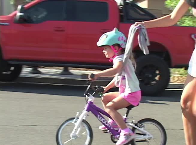 A mother helps her daughter learn to ride a bike down a residential street on a sunny day