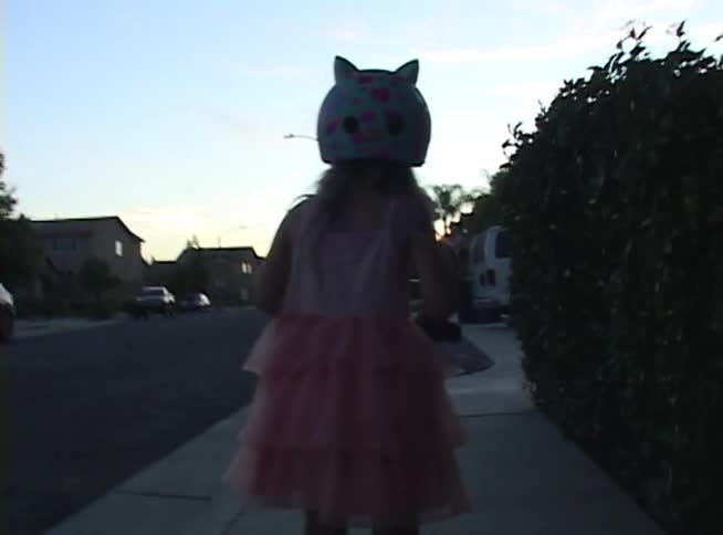 Nostalgic home video footage shows a little girl in a pink dress and cat helmet scootering down the sidewalk at twilight