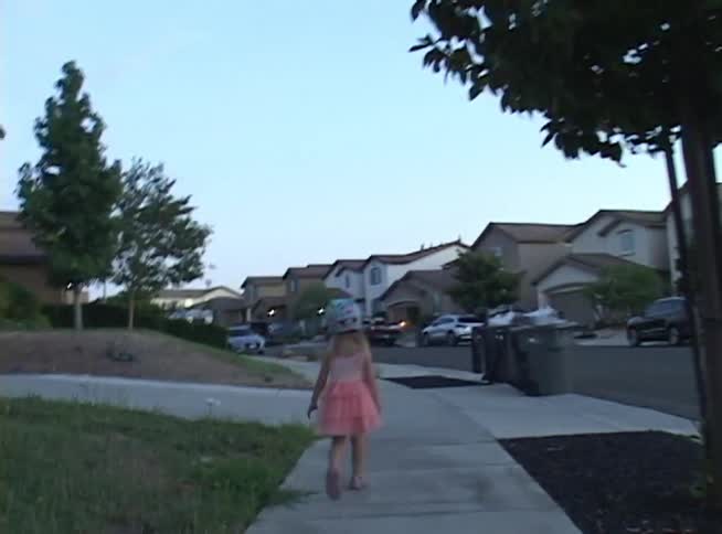 Rear view of a little girl in a pink frilly dress and a fun helmet walking in slow motion along the sidewalk in a quiet suburban neighborhood