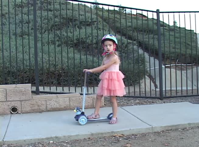 A cute little girl in a pink dress and helmet rides her scooter past a no trespassing sign on a suburban sidewalk
