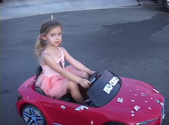 A patriotic little girl in a pink dress sits in her red toy convertible car on a suburban street