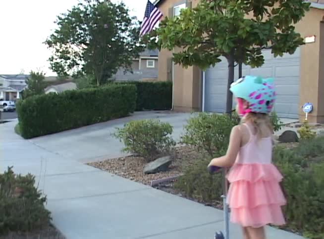 A young girl in a pink dress and a colorful helmet rides her scooter down a sidewalk in a suburban neighborhood
