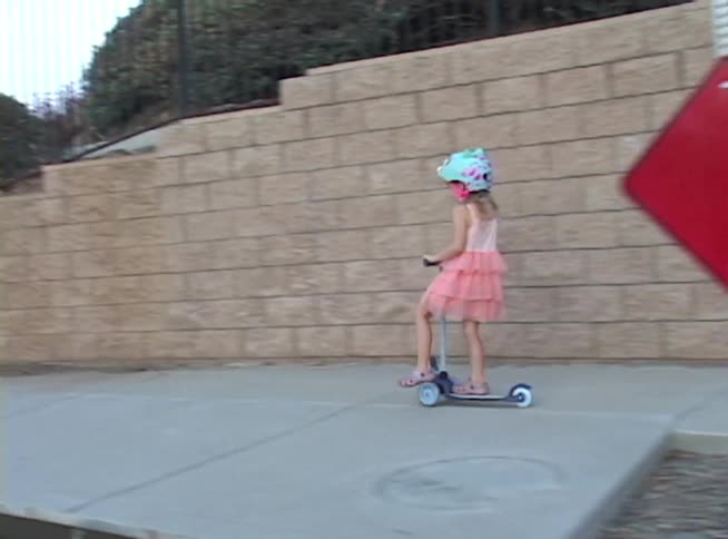 A young girl in a pink tutu dress and helmet rides her scooter in slow motion along a sidewalk