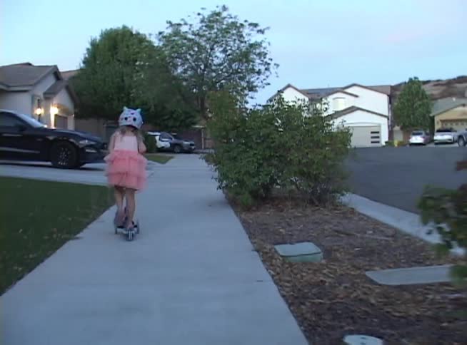 Following a young girl in a pink dress and a cute helmet as she rides her scooter down a suburban sidewalk at twilight
