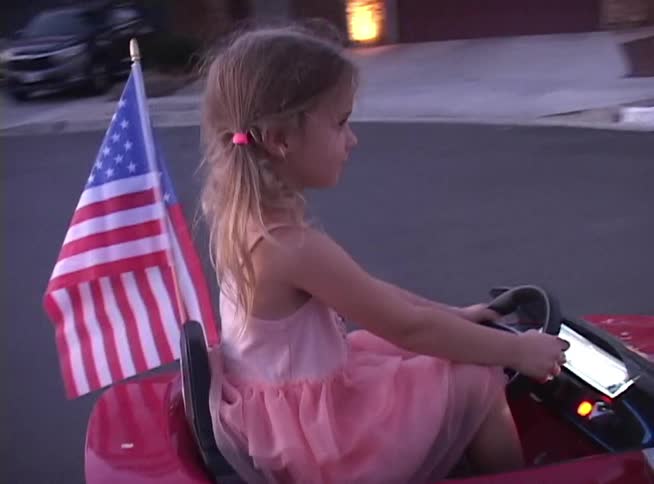 A cute young girl with pigtails joyfully waves as she drives her red toy convertible with an American flag down a suburban street at dusk