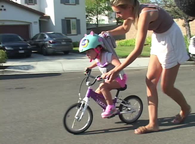 A mother patiently walks alongside her daughter, holding on to help her balance as the little girl learns to ride her bike on a sunny suburban street