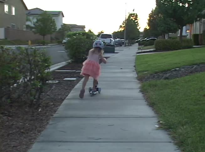 A young girl in a pink dress and a colorful cat-eared helmet rides her scooter along a sidewalk in a suburban neighborhood at dusk