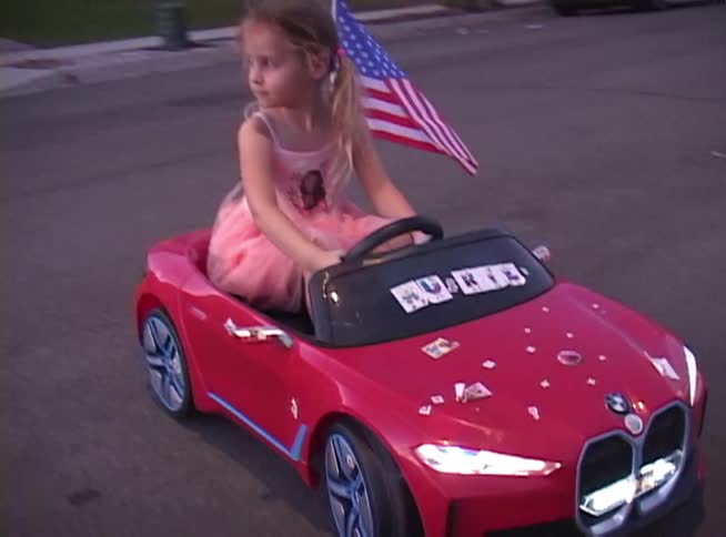 A cute young girl with pigtails patriotically drives her red toy convertible with an American flag down the street