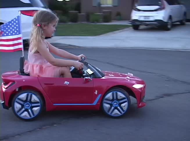 A patriotic little girl drives her red toy convertible car with an American flag through her suburban neighborhood at dusk
