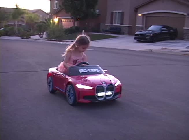 A young girl proudly drives her red electric toy car with an American flag down a quiet suburban street at dusk