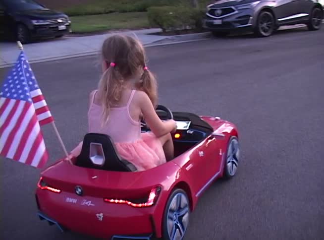 A cute little girl with pigtails drives her red toy BMW sports car with an American flag down a quiet suburban street