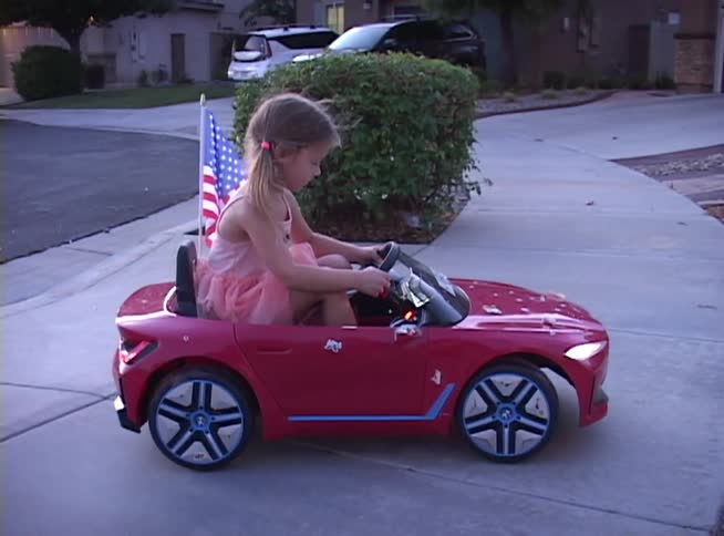A little girl in a pink dress proudly drives her red toy convertible with an American flag through her suburban neighborhood at dusk