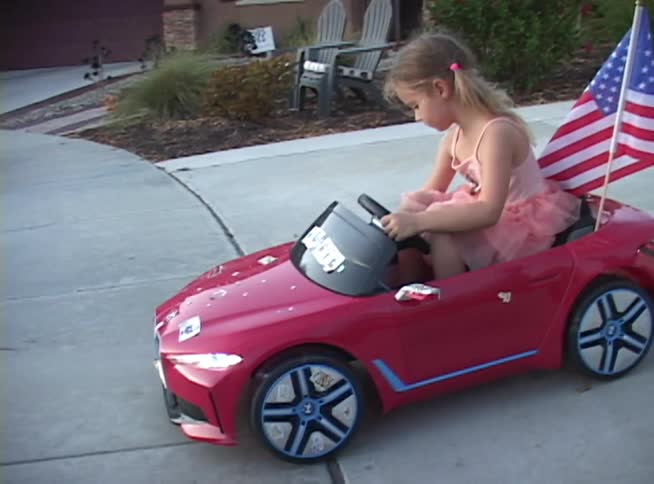 A cute little girl in a pink dress proudly drives her red toy convertible with an American flag down a suburban sidewalk