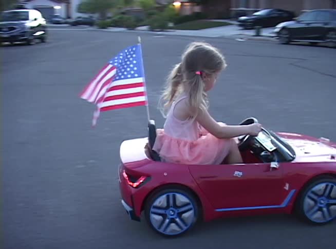 A cute little girl in a pink dress happily drives her red toy BMW car with an American flag down a suburban street
