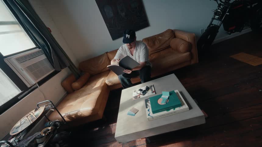A wide, high-angle view of a man in a baseball cap sitting on a leather sofa and reading a book in his sunlit urban loft