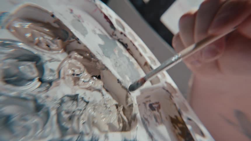 Super close-up slow-motion shot of an artist's hands mixing grey paint on a palette with a brush