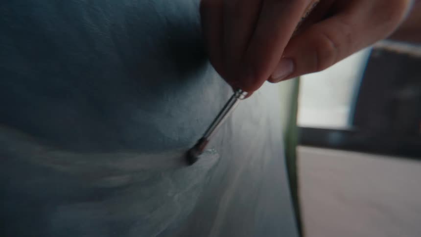 An artist's hand delicately paints a dark, swirling circle onto a canvas with a fine-tipped brush in a well-lit studio
