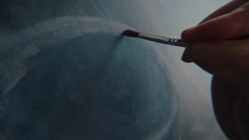 An artist gently applies strokes of paint with a fine brush to a blue abstract canvas in a slow-motion close-up
