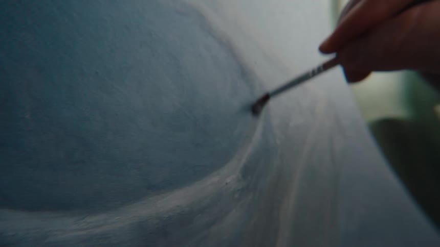 An artist's hand carefully applies paint with a fine brush to a blue canvas in a slow-motion close-up