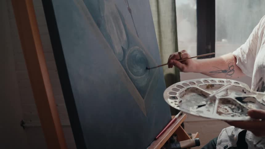 A close-up of a tattooed artist carefully adding detail to an abstract painting on an easel in their studio