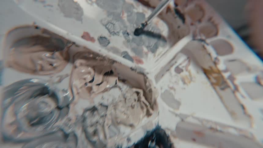 Close up of an artist's hand mixing dark paint colors on a palette with a brush in slow motion