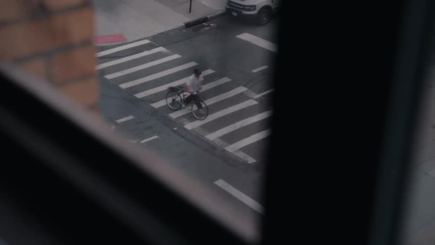 A slow-motion, high-angle view from a window shows a man on a bicycle riding through a crosswalk on a city street