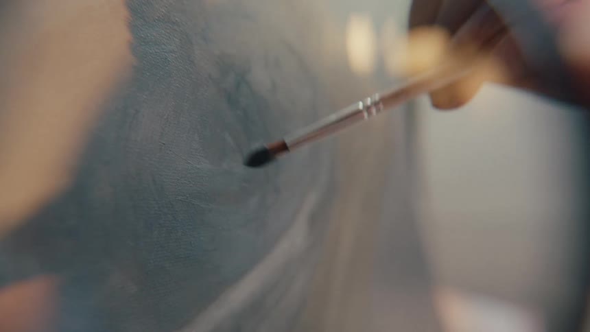A close-up shot shows an artist's hand using a paintbrush to apply strokes of grey paint onto a canvas