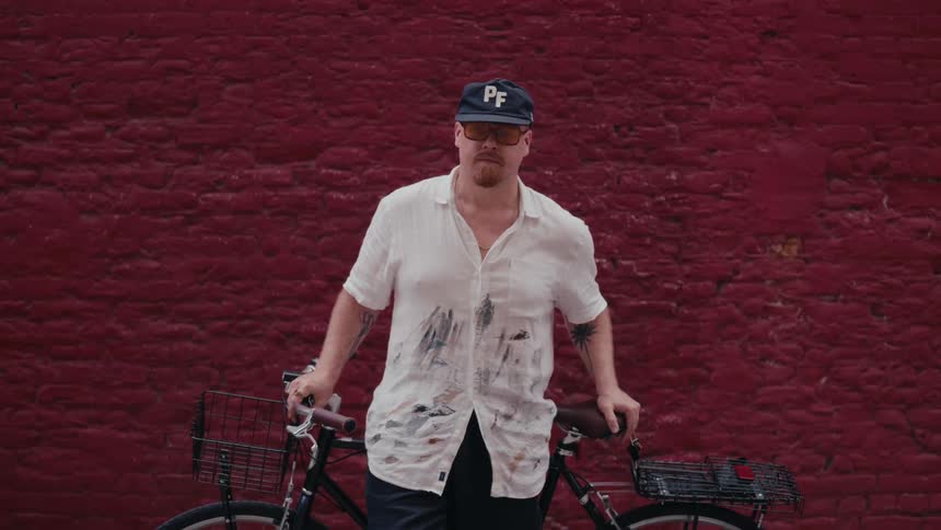 Portrait of a man in a paint-splattered shirt holding his bicycle against a vibrant red wall