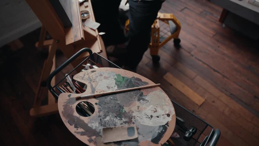 High-angle view of a painter's messy palette on a cart while the artist works at an easel in their studio