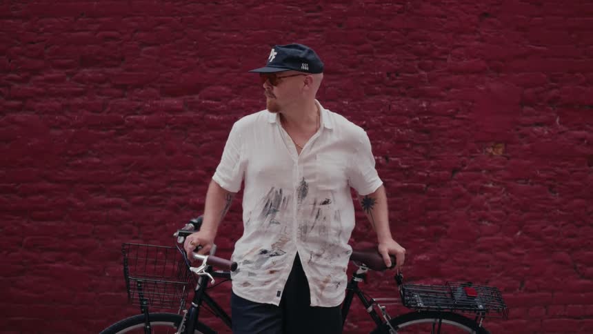 A stylish artist in a paint-stained shirt stands confidently with his bicycle against a red brick wall as another cyclist blurs past in the city
