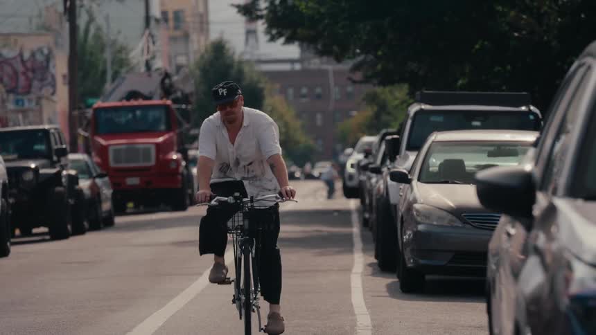A man in a baseball cap and sunglasses rides his bicycle in slow motion down an urban street lined with parked cars