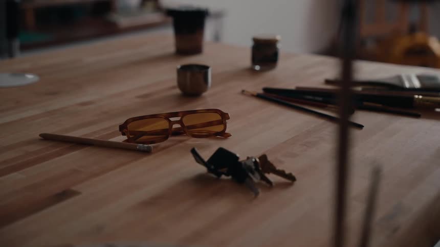 A hand places a set of keys on a wooden table alongside sunglasses and paint brushes