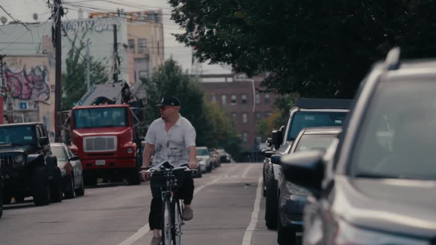 A man in a baseball cap and sunglasses cycles in slow motion down a gritty urban street lined with parked cars