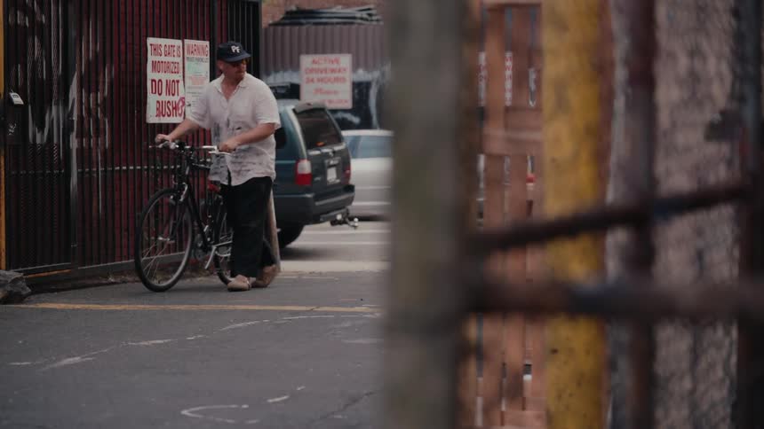 A man wearing a baseball cap walks his bicycle out of a gated urban alleyway