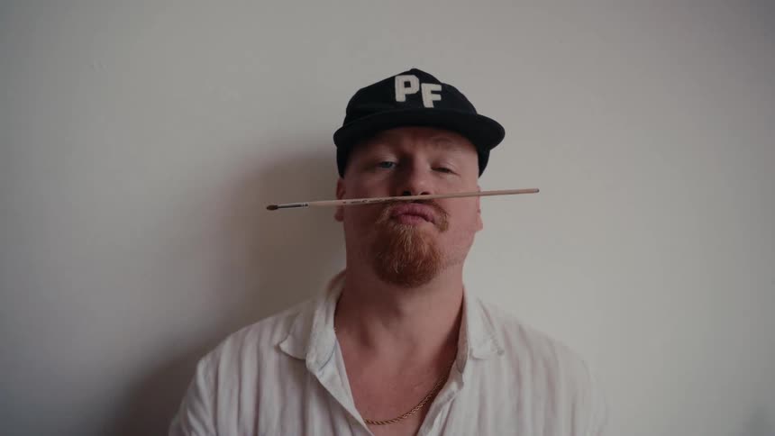 A quirky male artist with a red beard playfully balances a paintbrush on his upper lip like a mustache