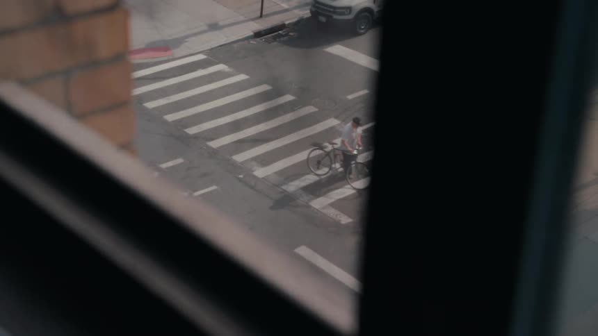 A high-angle view from a window captures a man riding his bicycle across a city street crosswalk