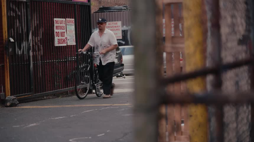 A man wearing a baseball cap slowly walks his bicycle out of a red, gated urban alleyway